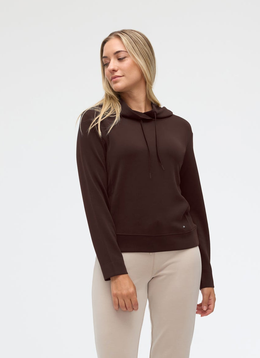 A woman modeling a dark brown long-sleeve hoodie and light beige jogger pants with white sneakers, standing against a plain white studio background.